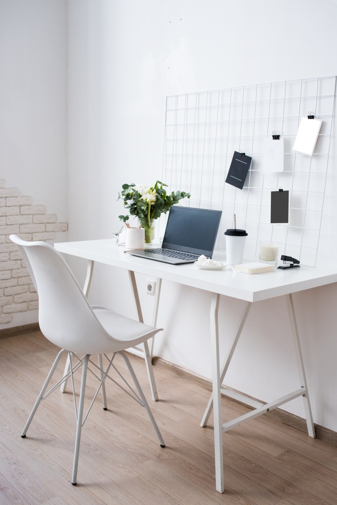 white minimalist workspace, white chair and simple white desk with laptop, plant, coffee cup and papers