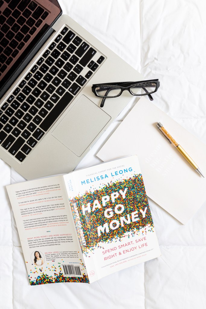overhead shot of a personal finance book - Happy Go Money, and a laptop, notebook and glasses
